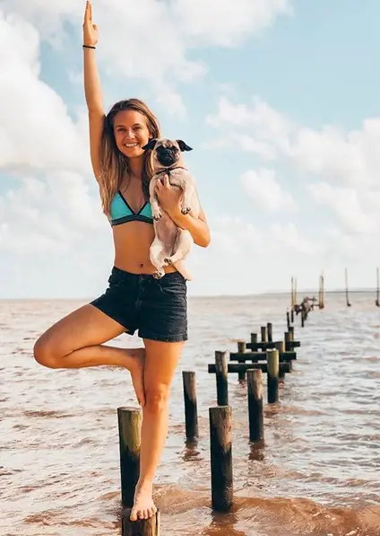 A woman doing yoga at the beach while carrying her Pug