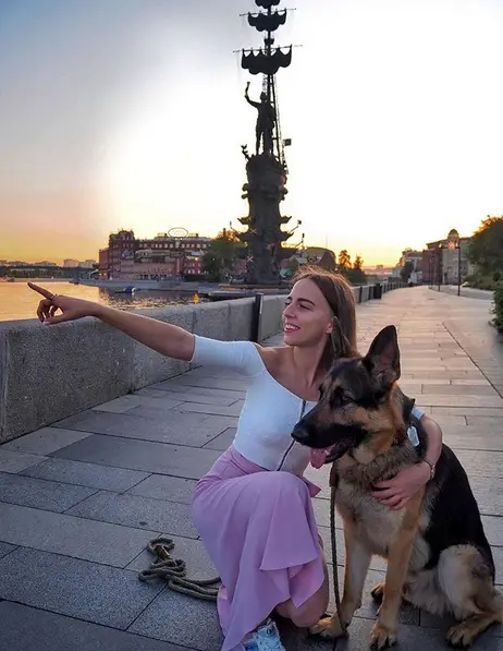 A woman kneeling on the pavement while pointing something in the sky while embracing a German Shepherd sitting next to her