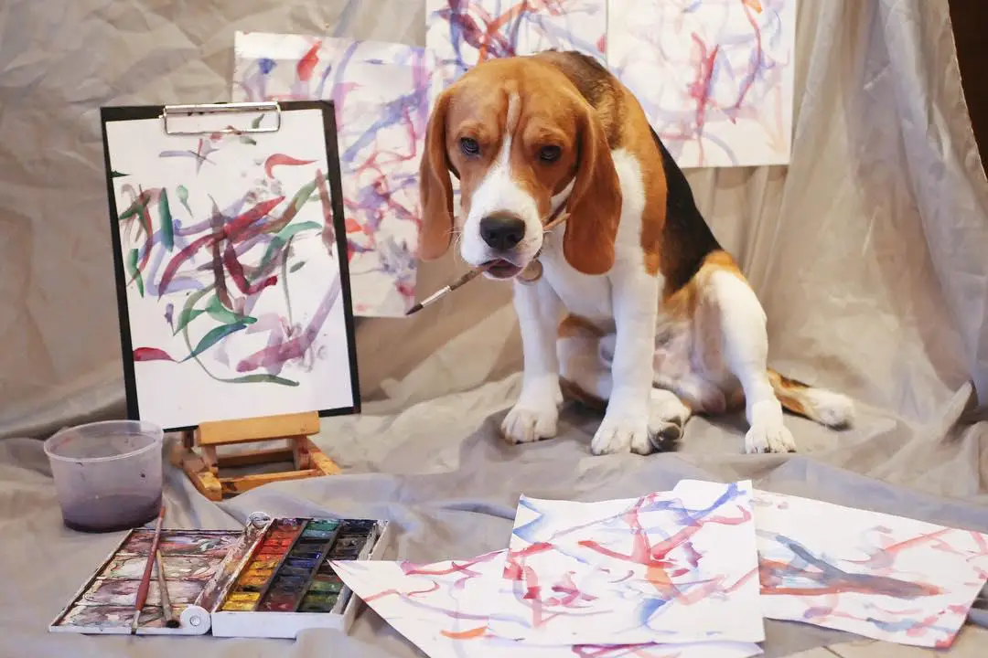 Beagle with a paint brush in its mouth while sitting on a fabric while being surrounded with paintings and palette