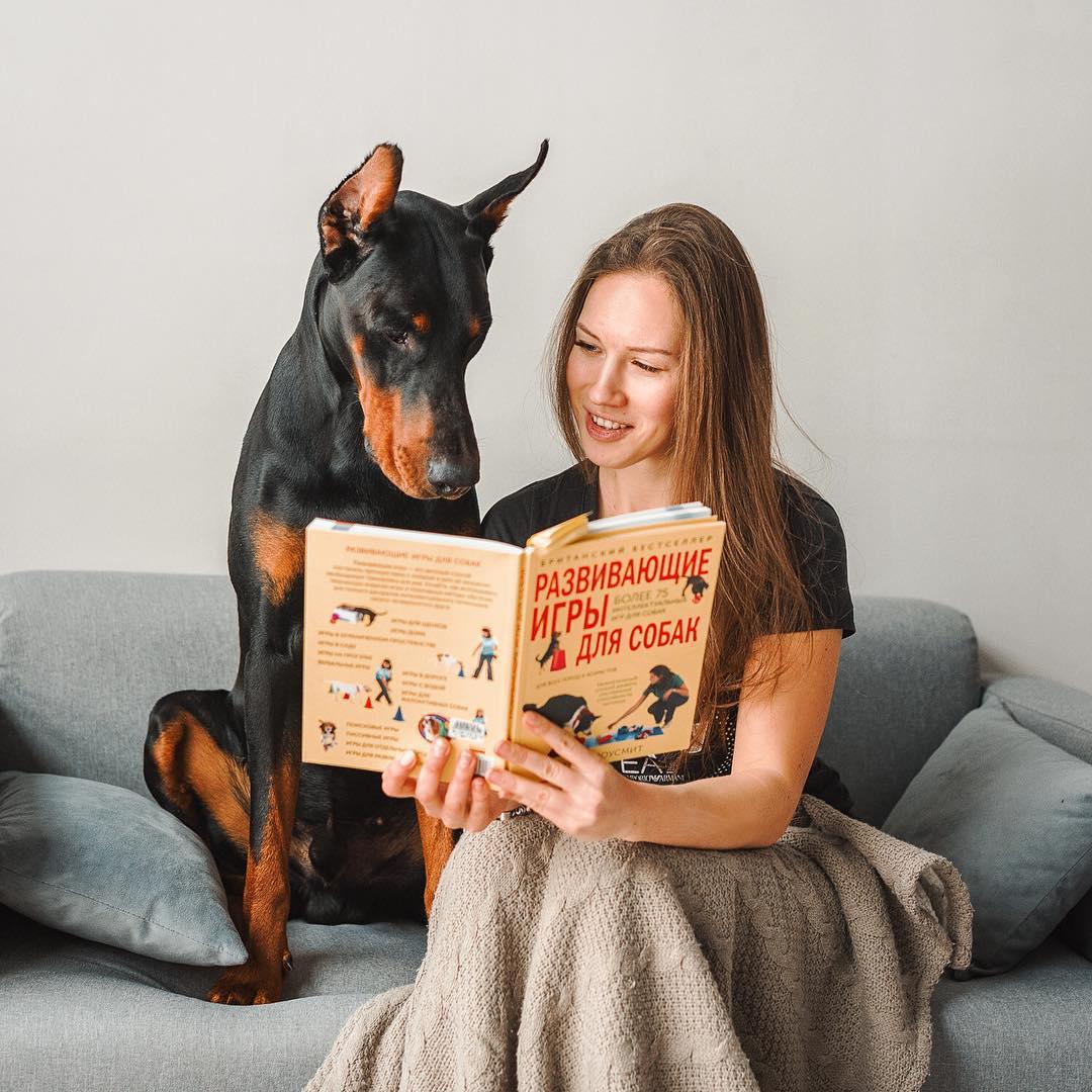 A woman on the couch reading a book to her Doberman sitting next to her