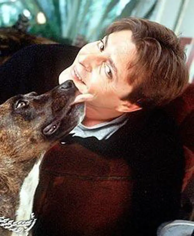 Michael J. Fox sitting on the couch while his cheeks is being licked by his pitbull