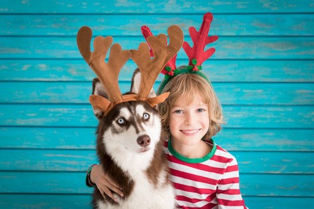 A kid wearing a reindeer headband while embracing and sitting next to a Husky also wearing reindeer headpiece