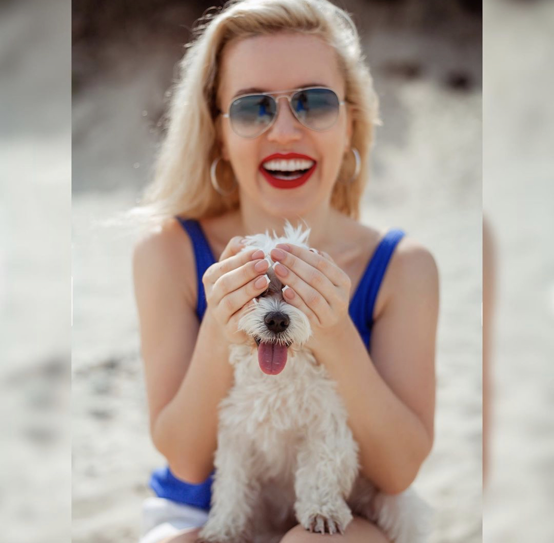 A woman sitting in the sand while covering the eyes of a Maltese sitting on top of her lap