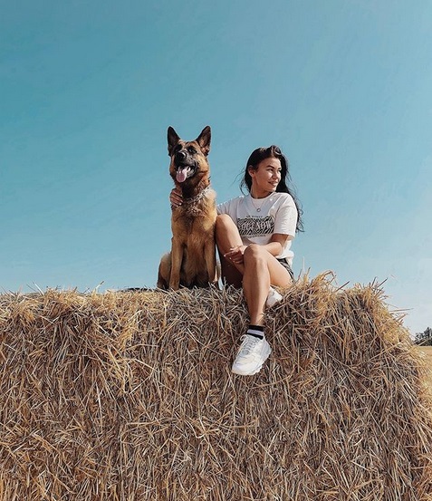 A German Shepherd woman sitting on top of the large bale of hay with her German Shepherd beside her