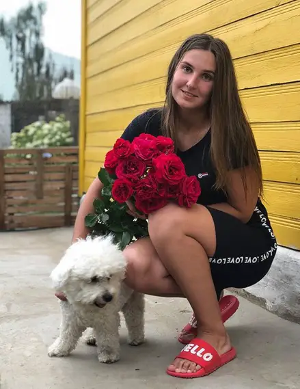 a woman with a bouquet of red roses beside her Bichon Frise standing on the floor
