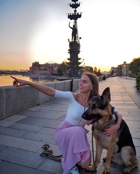 A German Shepherd sitting on the pavement next to a woman pointing at something