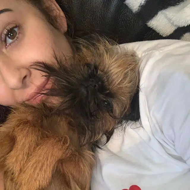 A woman taking a selfie with her Brussels Griffon while lying next to each other on the bed