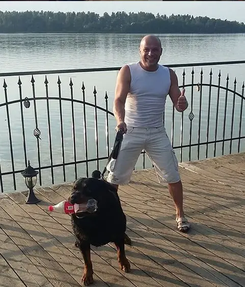 A man standing behind the railing by the ocean while holding the leash of a Rottweiler holding an empty plastic soda in its mouth
