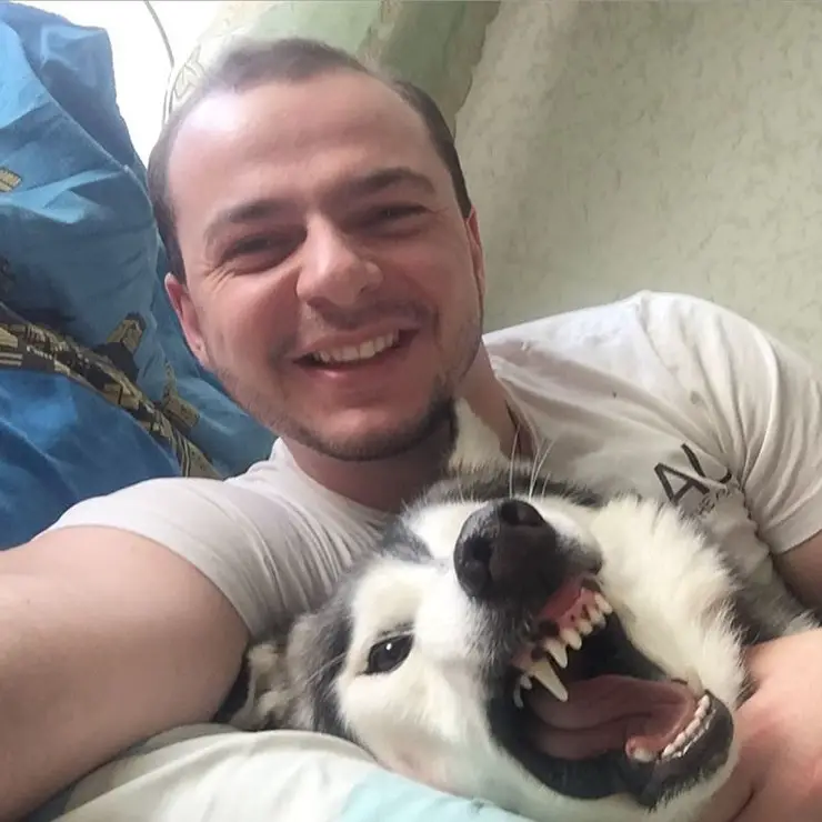 A man lying on the bed while taking a selfie next to a Husky with its mouth wide open