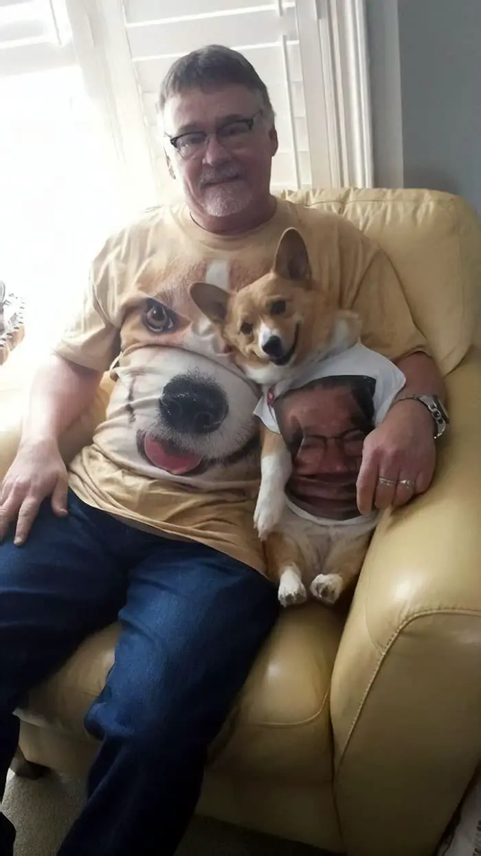 A man sitting on the chair wearing a shirt with the face of the corgi sitting beside him and wearing a short printed with his face