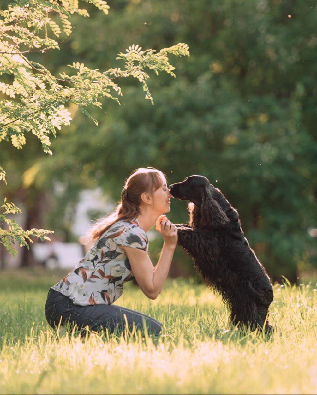 A woman kneeling in the grass with her black Cocker Spaniel leaning toward her and licking her nose