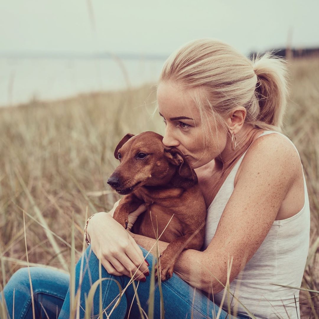 A woman sitting in the field of grass while kissing her Miniature Pinscher lying on her lap