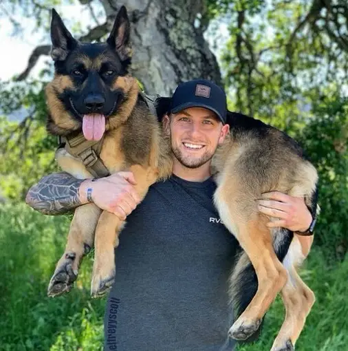 a man standing under the tree while carrying a German Shepherd over his shoulder