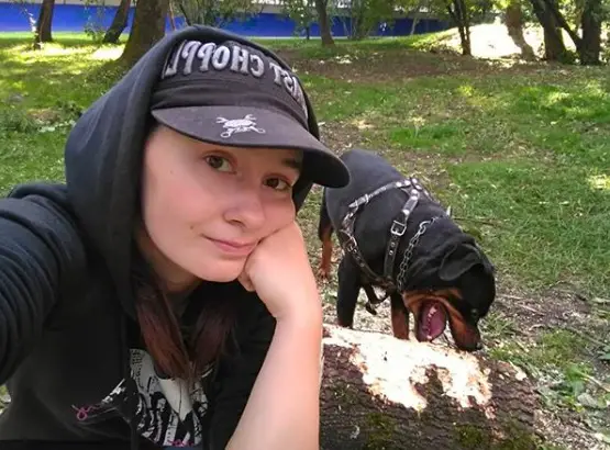 A woman taking a selfie with her Rottweiler biting the laid tree trunk at the park